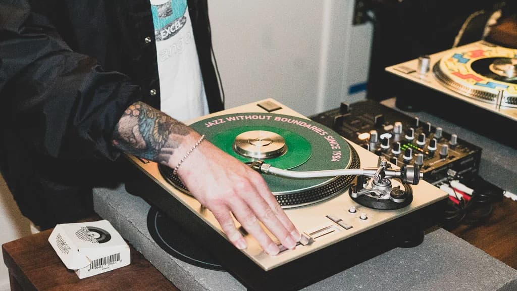 A DJ's tattooed hand on a turntable, a green 'Jazz Without Boundaries Since 1984' record on the platter.
